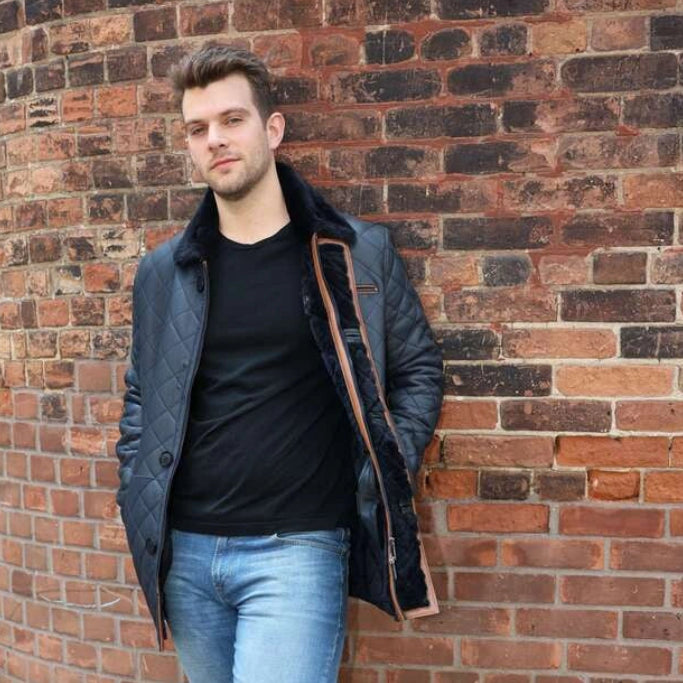 Man leaning against a brick wall wearing a coat, black shirt, and jeans.