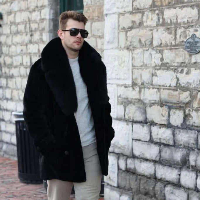 Man in a black coat and sunglasses standing against a stone wall.