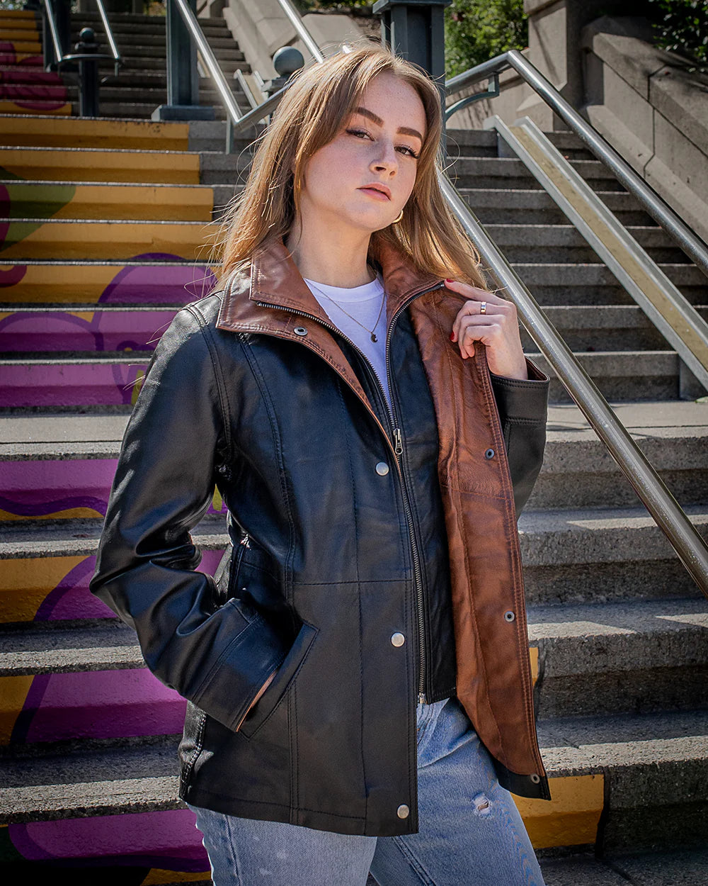 Lifestyle photoshoot of a woman wearing the Adeline black leather car coat, showcasing its elegant 3/4 length and regular fit  with a brown leather collar on a colorful staircase.