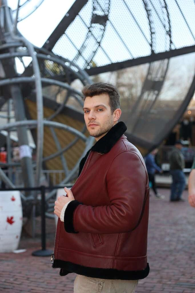 Man wearing Aurelius Burgundy B3 Bomber Jacket with black fur trim standing in front of a large metal sculpture.