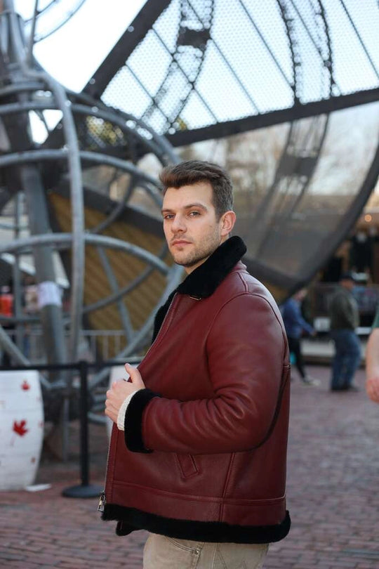 Man wearing Aurelius Burgundy B3 Bomber Jacket with black fur trim standing in front of a large metal sculpture.