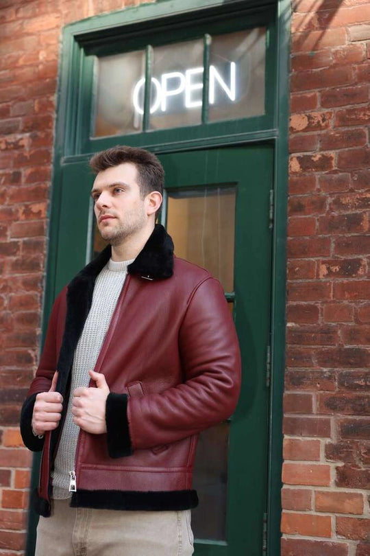 Man wearing Aurelius Burgundy B3 Bomber Jacket  with fur trim standing in front of a brick wall and open door.