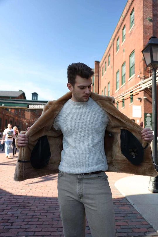 Man wearing a light blue sweater and beige pants with Classic Ginger Brown B3 Bomber Aviator Shearling Jacket over his shoulder showcasing interior of the jacket, standing on a brick sidewalk.
