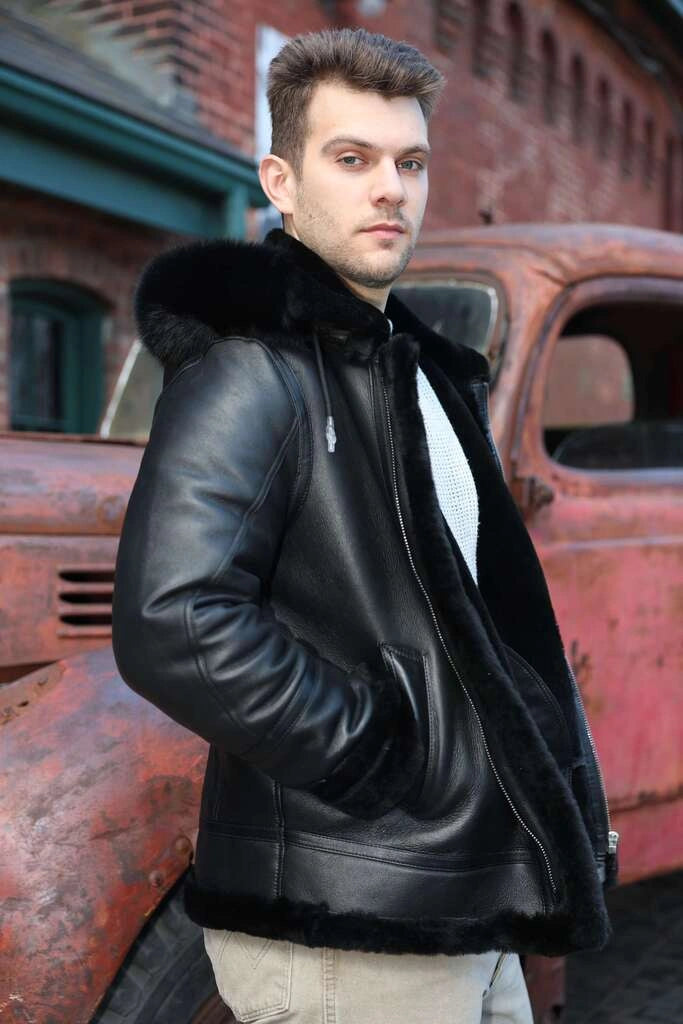 Man wearing Felix black bomber shearling jacket with fur trim standing next to an old, rusted vehicle.