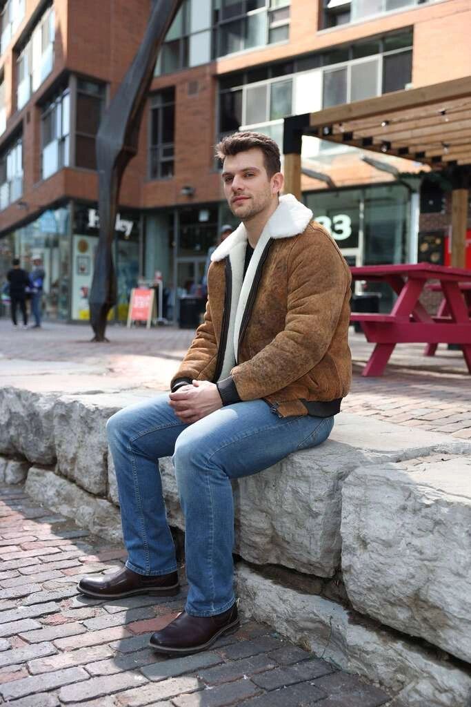 Man sitting on a stone bench wearing Lucian's distressed brown A2 Bomber shearling jacket in an urban setting.