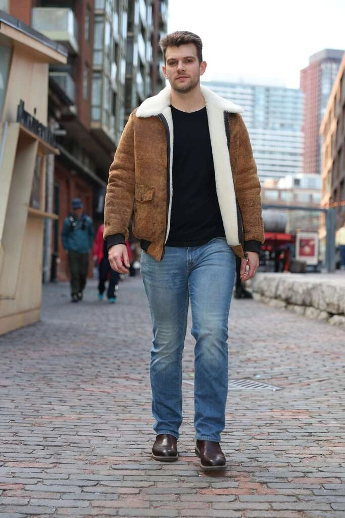 Man walking on a cobblestone street wearing a Lucian's distressed brown A2 Bomber shearling jacket, black shirt, and blue jeans.