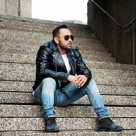 A male model wearing sunglasses and the Perfect Biker Style heavy leather biker jacket, sitting on concrete steps that highlights the asymmetrical zipper and belted waist.