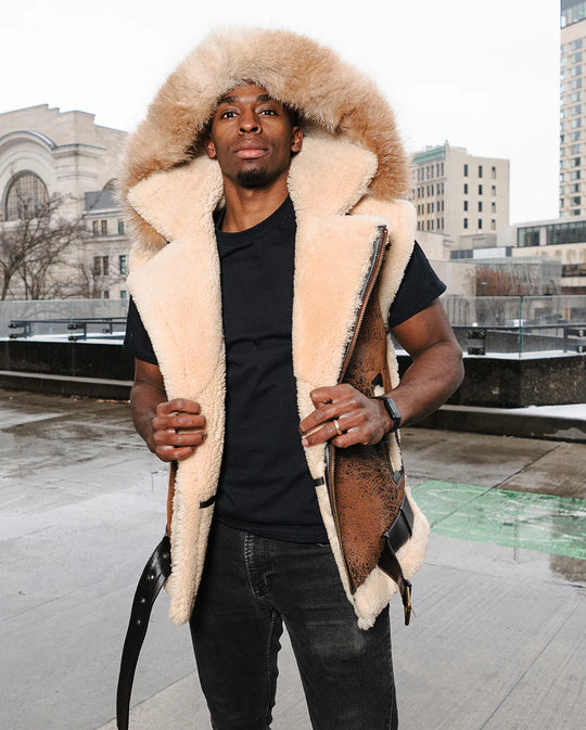Lifestyle image of a model standing in Ridgeway's Vintage Shearling Vest, featuring rugged brown leather and fluffy shearling lining. The model pairs the vest with a black t-shirt and jeans, exuding a cool, casual style outdoors.